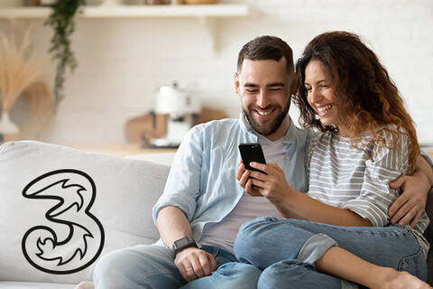 glückliches junges Paar auf Sofa schaut zusammen auf Smartphone neben Drei Logo
