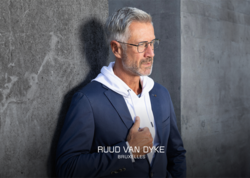 Ein Mann in sportlich elegantem Outfit lehnt an einer Mauer und blickt in die Ferne. Er trägt eine Brille der Marke Ruud van Dyke. Auf der Grafik steht folgender Text: "Ruud van Dyke Bruxelles"