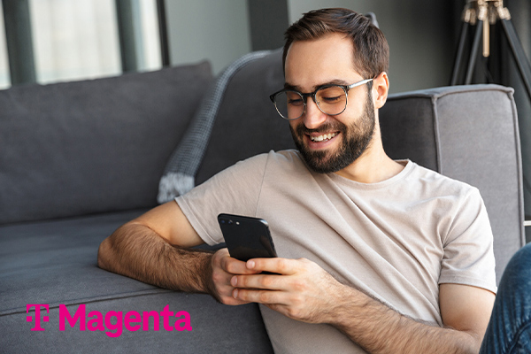 Mann mit Smartphone lehnt sitzend an Couch neben Magenta-Logo