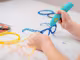 Auf der Grafik sind Kinderhände zu sehen, die einen 3D-Stift halten. Damit wird gerade eine blaue Brille angefertigt. Rundherum befindet sich das Zubehör sowie eine gelbe Brille.
