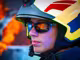 Feuerwehrmann mit einem Helm und einer Sonnenbrille von Hartlauer, verschwommenes Feuer im Hintergrund