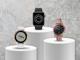 drei verschiedene Smartwatch-Modelle auf einem Podest vor grauem Zahnrad-Hintergrund