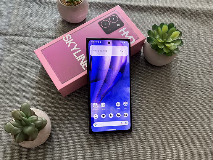 Das HMD Skyline liegt halb auf der Verpackung des Ger&auml;ts in der Farbe neon pink. Der Bildschirm ist eingeschaltet und entsperrt. Darunter befindet sich ein Leinentuch und rundherum sind drei Pflanzen platziert.