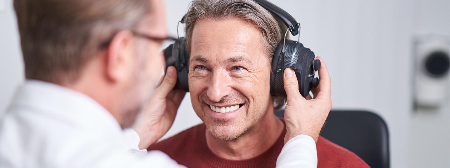 Hartlauer Hörakustiker setzt einem lächelnden Kunden Kopfhörer auf 