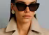Eine Frau in einem braunen Mantel trägt eine schwarze Sonnenbrille.