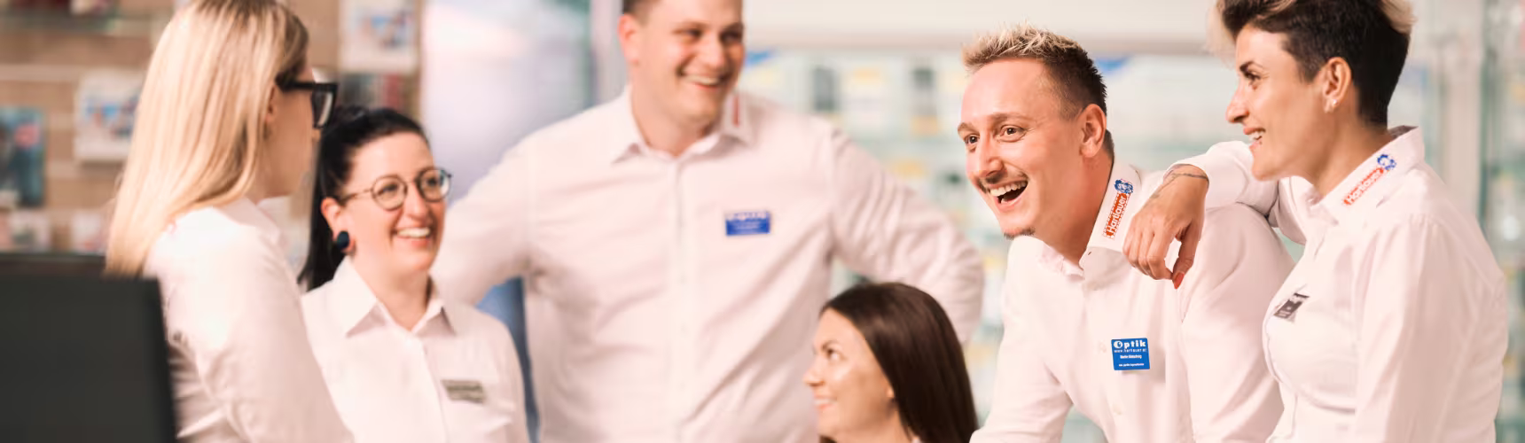 "Sechs Hartlauer Mitarbeiter und Mitarbeiterinnen stehen und sitzen in einem Kreis und lachen gemeinsam."