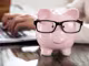 Ein Keramikschwein trägt eine schwarze Brille. Dahinter sind Hände auf einer Laptop-Tastatur zu sehen.