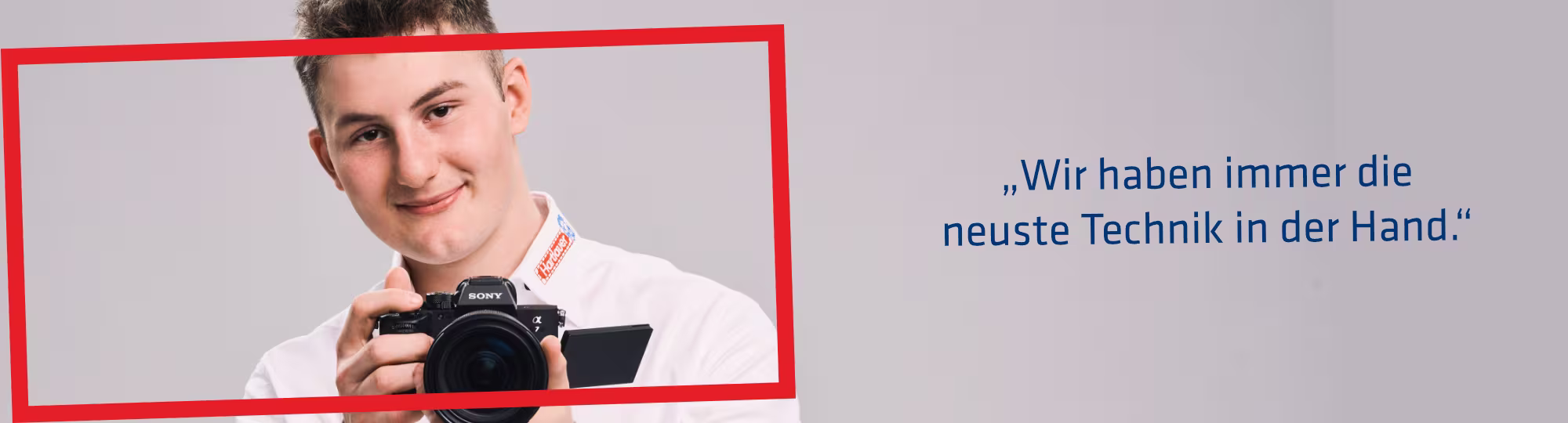 Ein Hartlauer Mitarbeiter hält eine Kamera in seinen Händen. Neben ihm befindet sich eine Sprechblase mit folgendem Text: "Wir haben immer die neueste Technik in der Hand." Die Grafik hat einen roten Rahmen.