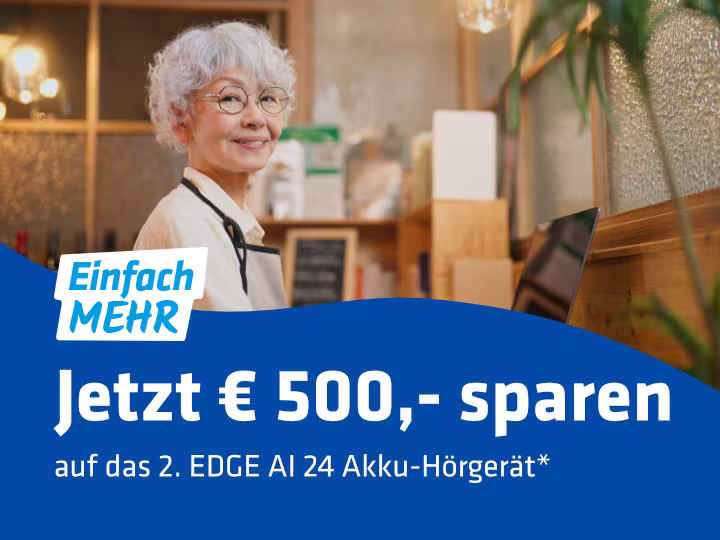 Im Bild ist eine Dame mit grauem kurzem Haar zu sehen. Sie trägt eine Brille. Sie steht gerade in einem Geschäft beim Laptop. Unten im Bild ist die blaue Welle mit dem Slogan "Jetzt € 500,- sparen auf das 2. EDGE AI 24 Akku-Hörgerät.*" Rechts zirka mittig und halb in der Welle ist das Logo "Einfach MEHR" zu sehen.